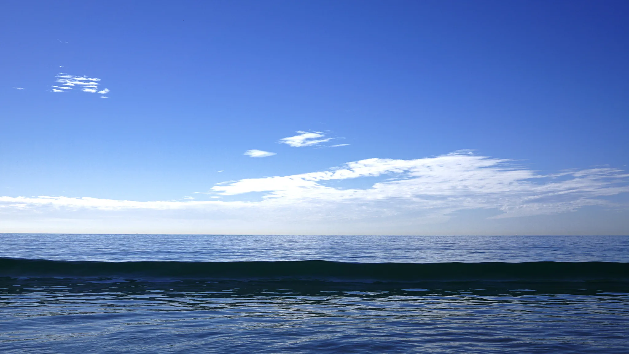 Dark ocean swell stretching across the foreground beneath a bright blue sky and thin horizon clouds