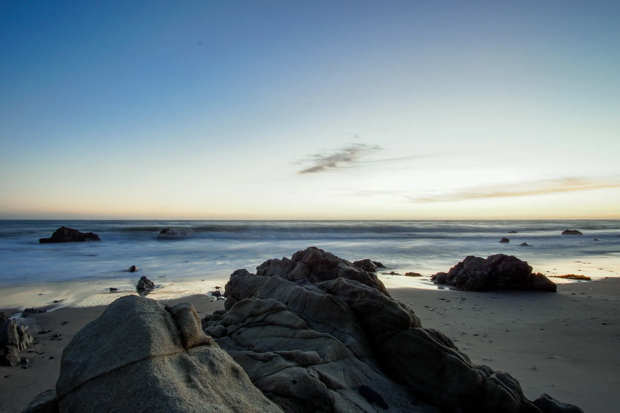 Dark coastal rocks and soft blurred surf under a pale sunset sky in Malibu