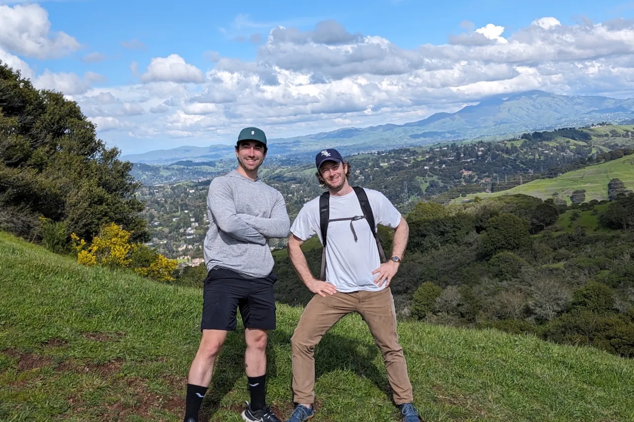 Two people hiking on a green hillside with mountains in the background