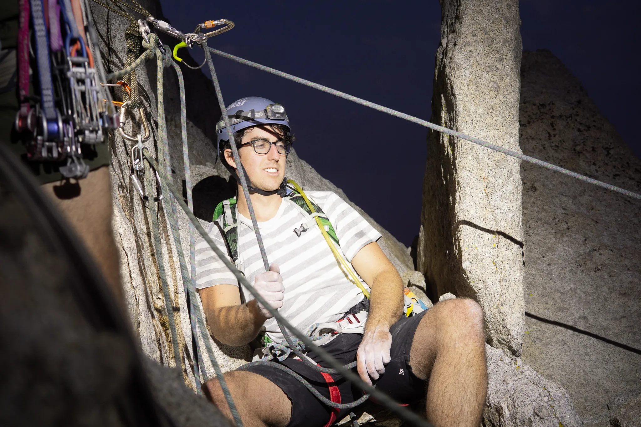 Climber in a helmet resting on a granite ledge at night, chalked hands and ropes clipped into the rock