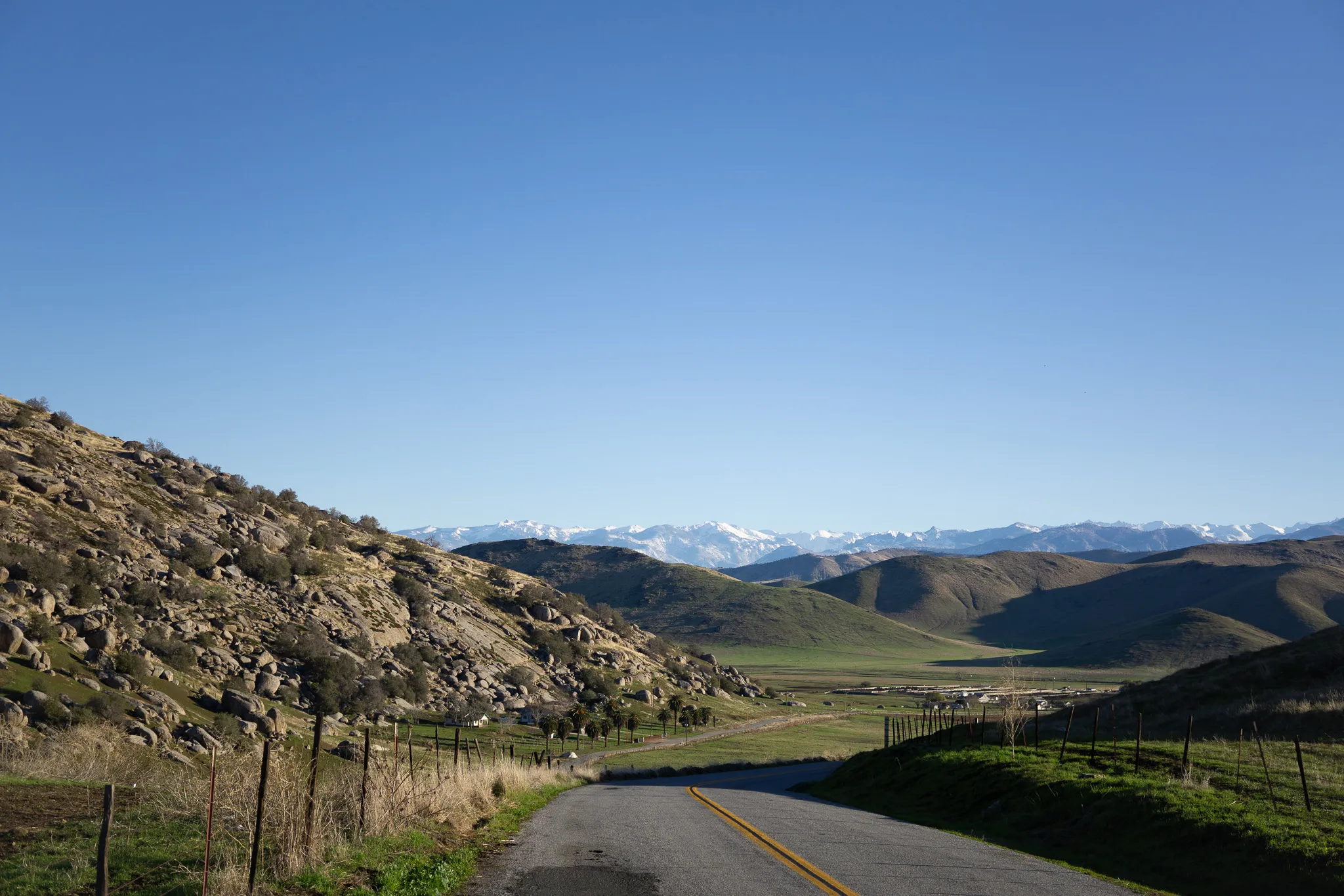 Two-lane road curving through rocky foothills toward a green valley and distant snowy mountains